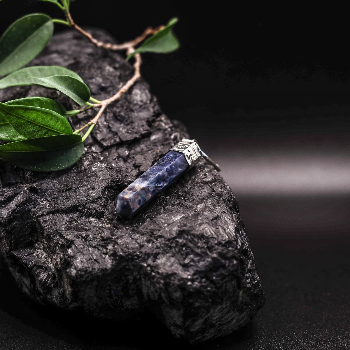 Djupblå lapis lazuli-hänge med guldiga pyritfläckar och silverfärgat mässingsfäste, omgiven av gröna växtblad