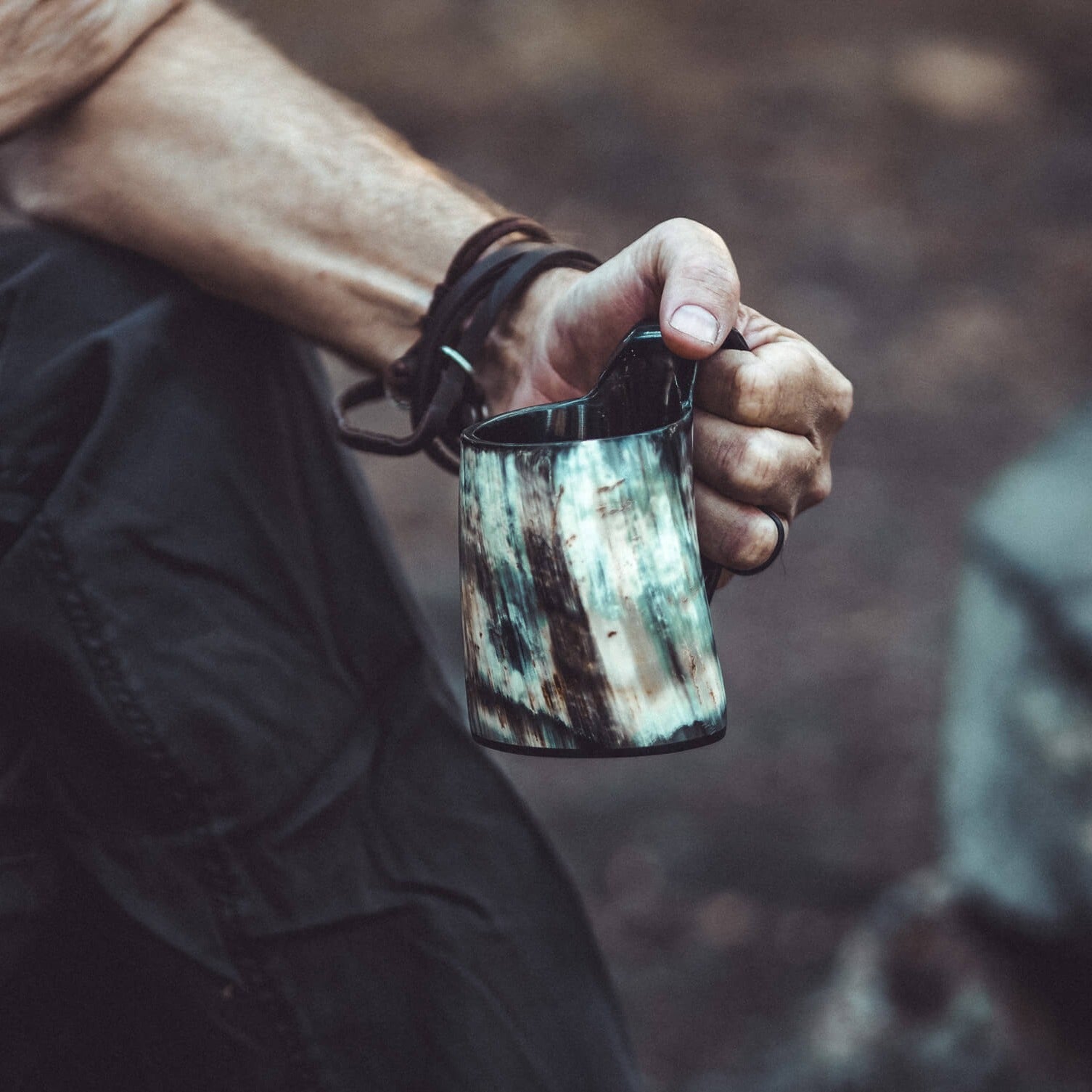 Närbild på mörk hornmugg i hand med läderarmband synligt. Visar muggens storlek och hornets naturliga karaktär.