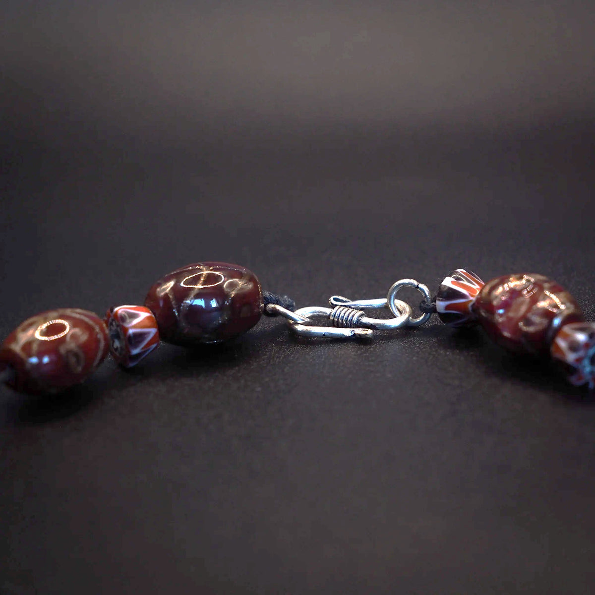 Närbild av S-format halsbandslås i silver kopplat till dekorerade glaspärlor mot mörk bakgrund