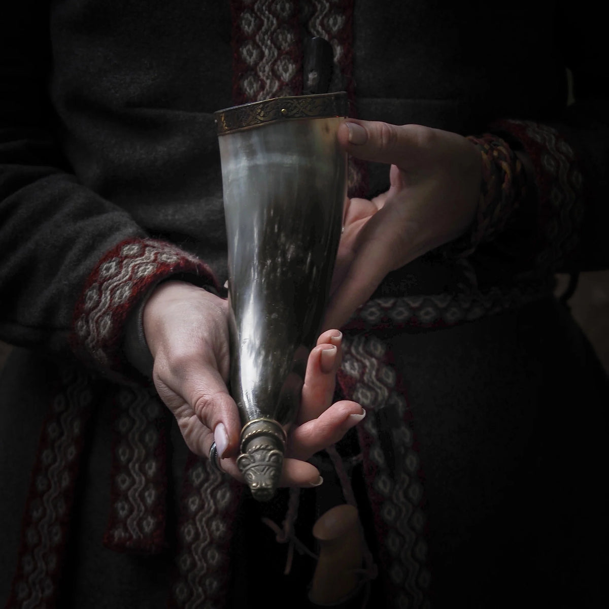 Hornmugg hållen i händer iförd vikingainspirerad väv, visande polerad horn med mässingdetaljer och traditionellt hantverk