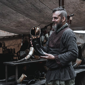 Stort dryckeshorn på cirka 4-5 liter hållet av vikingatida hantverkare i verkstad. Mässingsband vid öppningen, naturlig mörkbrun hornfärg, autentisk historisk miljö synlig i bakgrunden.