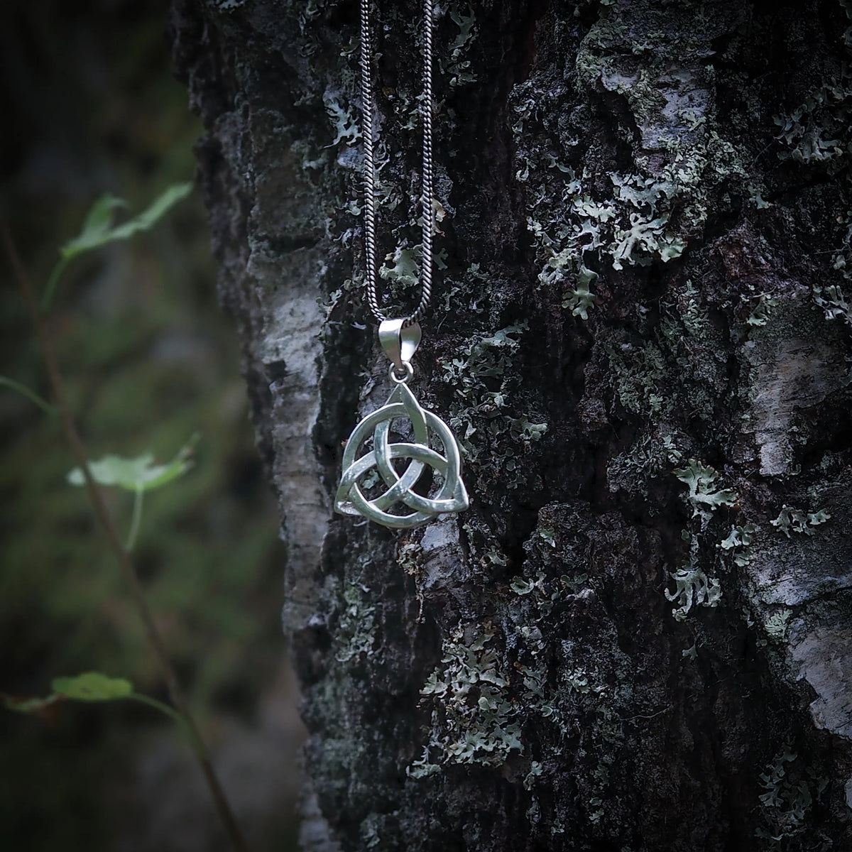 Silverbelagd evighetsknut-hängsmycke på en metallkedja hängande från trädstammen, fotograferad i närbild mot naturbakgrund