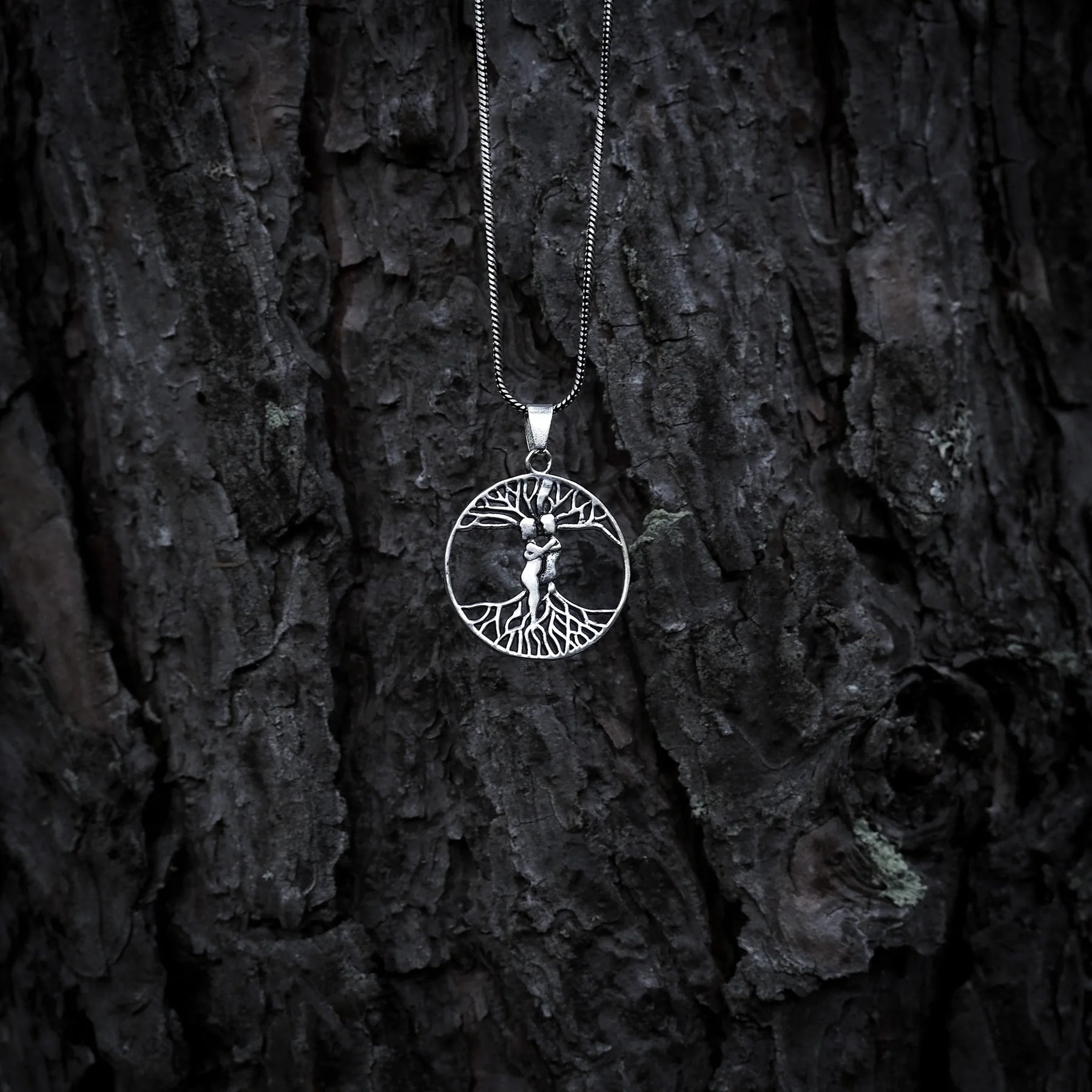 Silverkedja med cirkelformad amulett föreställande Yggdrasil, världsträdet, med synliga rötter och grenar på mörkrändig bakgrund.