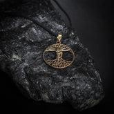 Ask och Embla-amulett i mässingsfärg med detaljerad Yggdrasil-träd design fotograferad på mörk sten