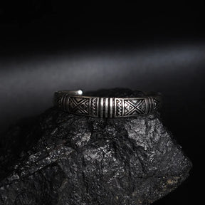En silverfärgad mässingsarmband med vikinginspirerade mönster av trianglar och linjer, fotograferad på mörk stenyta.