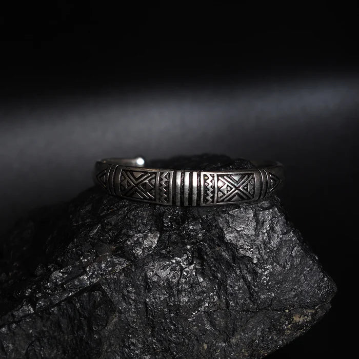 En silverfärgad mässingsarmband med vikinginspirerade mönster av trianglar och linjer, fotograferad på mörk stenyta.
