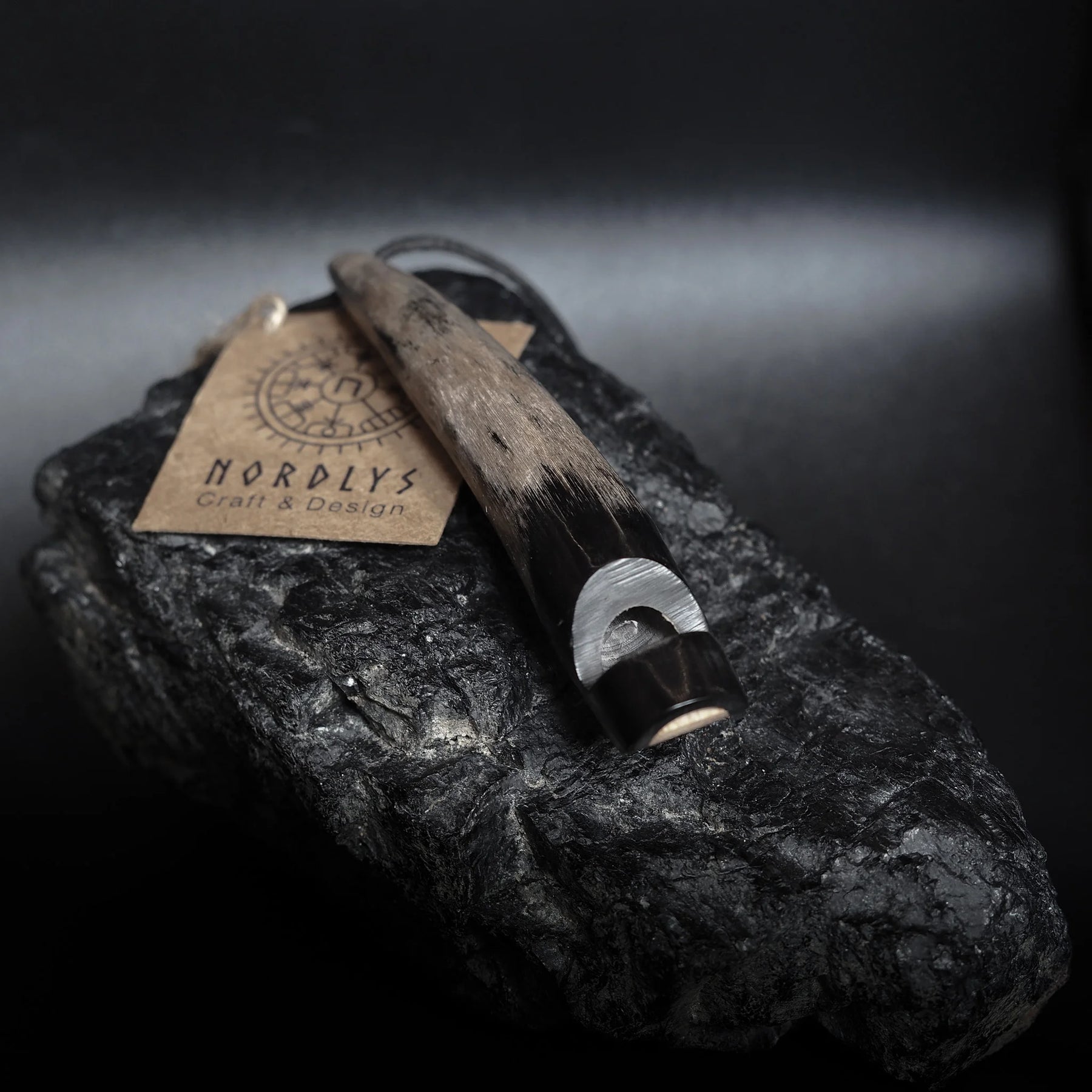 Visselpipa av horn med svart lädersnöre och Nordlys Craft & Design-etikett. Mörkhorn med synliga kornmönster på sliten stenyta. Professionell produktfotografi som visar det handgjorda detaljarbetet.