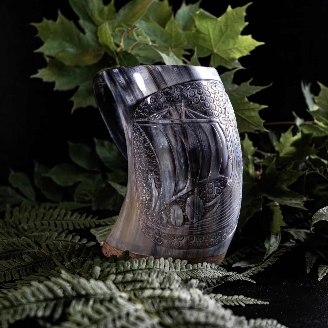 Handsnidad hornmugg med vikingskepp-gravyr omgiven av ormbunks- och ekblad, visar intrikat traditionell utskärning av långskepp-design.