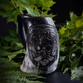 Slutet porträtt av Lokemask på hornmugg. Kraftfull skuren ansiktesuttryck med framträdande ögonhålor och stiliserad skäggmönster. Mässingsdetaljer skapar kontrast med punkterad textur. Mörk horn i naturlig belysning.