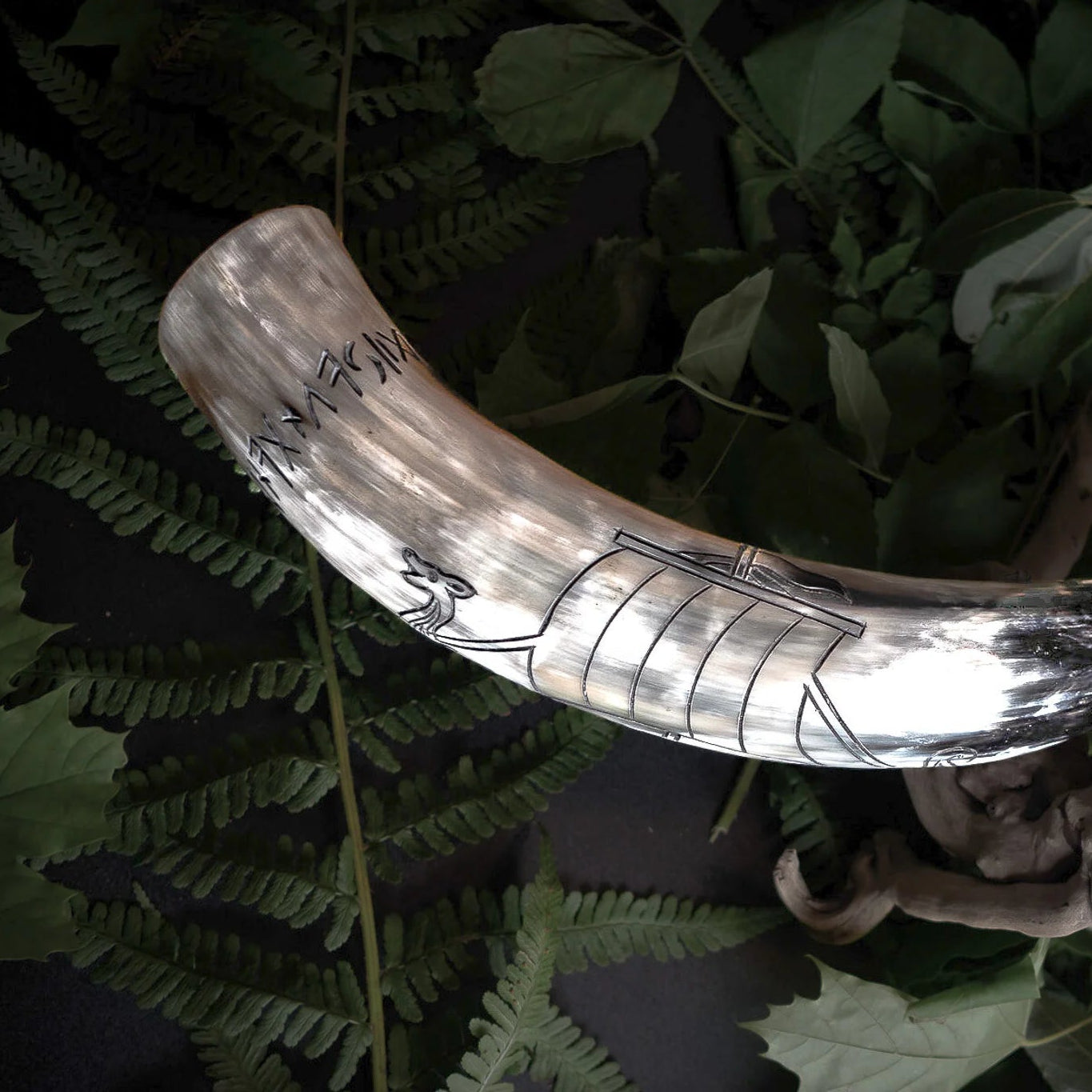 Naturligt tjurhorn med graverad Vikingaskepp och runinskripsioner. Hornet visar naturlig kremfärgning med detaljerade svarta graveringslinjer av Viking-långskeppet med årarmarkeringar och runor. Grön ormbunkbakgrund. Professionell studiobelysning framhäver graveringsdetaljer och naturlig hornstruktur.