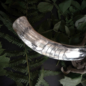 Naturligt tjurhorn med graverad Vikingaskepp och runinskripsioner. Hornet visar naturlig kremfärgning med detaljerade svarta graveringslinjer av Viking-långskeppet med årarmarkeringar och runor. Grön ormbunkbakgrund. Professionell studiobelysning framhäver graveringsdetaljer och naturlig hornstruktur.