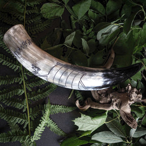 Naturligt horn med graverad Vikingaskepp och runor från Bergen, Norge. Hornet visar mörksvart färgning med röd/koppargul gravering av Viking-långskeppet med distinkiv framstäv, årmar och mast. Runskriftsinskripsioner löper längs skeppet. Fotograferat horisontellt på träverk.