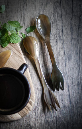 Två ljusa oxhorn-sporks presenterade vertikalt sida vid sida. Båda visar gaffol och skalkis-ändamål. Naturligt horn med kornmönster. Rent, katalog-lämpat produktfoto.