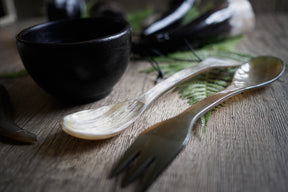 Tre ljusa oxhorn-sporks och mörkbrun keramik skål på trämatta. Sporkarna visar gaffol och skalkis, naturligt kornmönster synligt. Ormbunksbladsdekoration i bakgrunden. Studio-stil produktfoto.