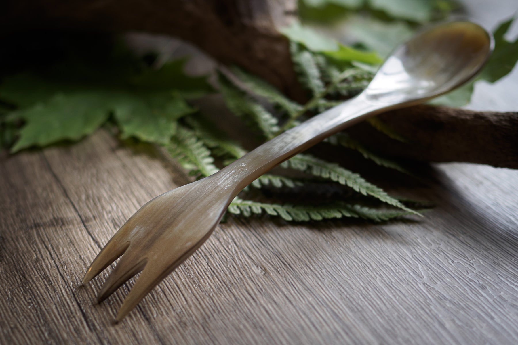 Oxhorn-spork i ljus färg, sida-profil. Gaffeln pekar upp åt höger, skeden åt vänster. Naturligt horn med synliga ådringar och kornstruktur. Grön ormbunksbladsdekoration på mörk träyta.