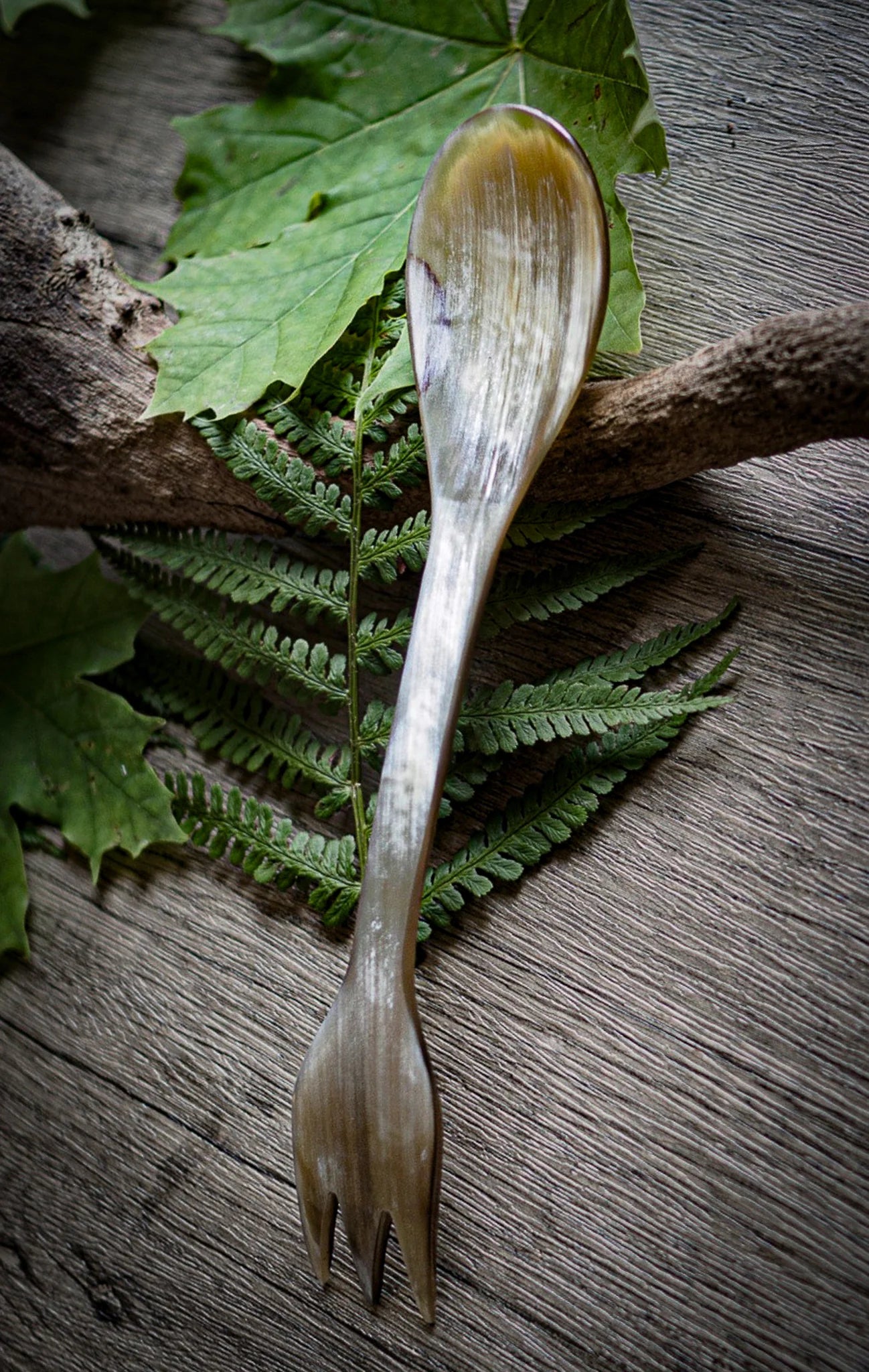 Spork i oxhorn fotograferad tillsammans med naturlig vegetation och trä för att framhäva miljövänlig camping-användning. Ljus ensfärgad hornyta, cirka 19 cm långt verktyg med skopa i ena änden och gaffel i den andra. 100% kompostbart.