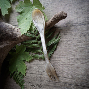 Spork i oxhorn fotograferad tillsammans med naturlig vegetation och trä för att framhäva miljövänlig camping-användning. Ljus ensfärgad hornyta, cirka 19 cm långt verktyg med skopa i ena änden och gaffel i den andra. 100% kompostbart.
