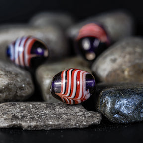 Blå pärla med vit-röda ränder. Tre pärlor fotograferade mot grå stenar. Pärlorna är rundade med horisontella vita och röda ränder. Material: glas. Användning: pärlor för smycken och smycken. Vikingainspirerad nordisk design.