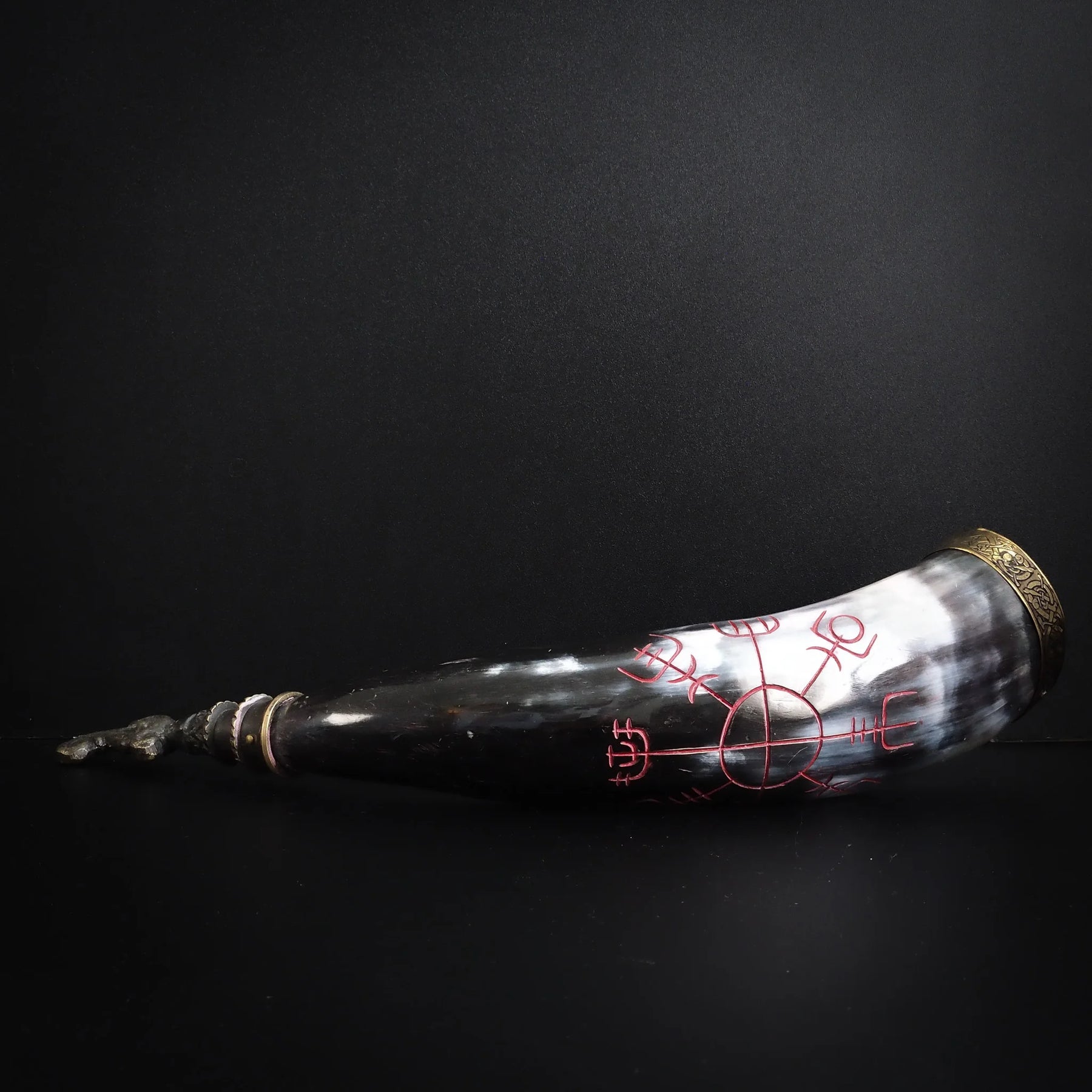 Graverad dryckeshorn med mörkt horn och roströda graveringar. Runsymboler och vikingamotiv utformade i cirkelformat mönster. Mässingsbåge synlig vid öppningen.