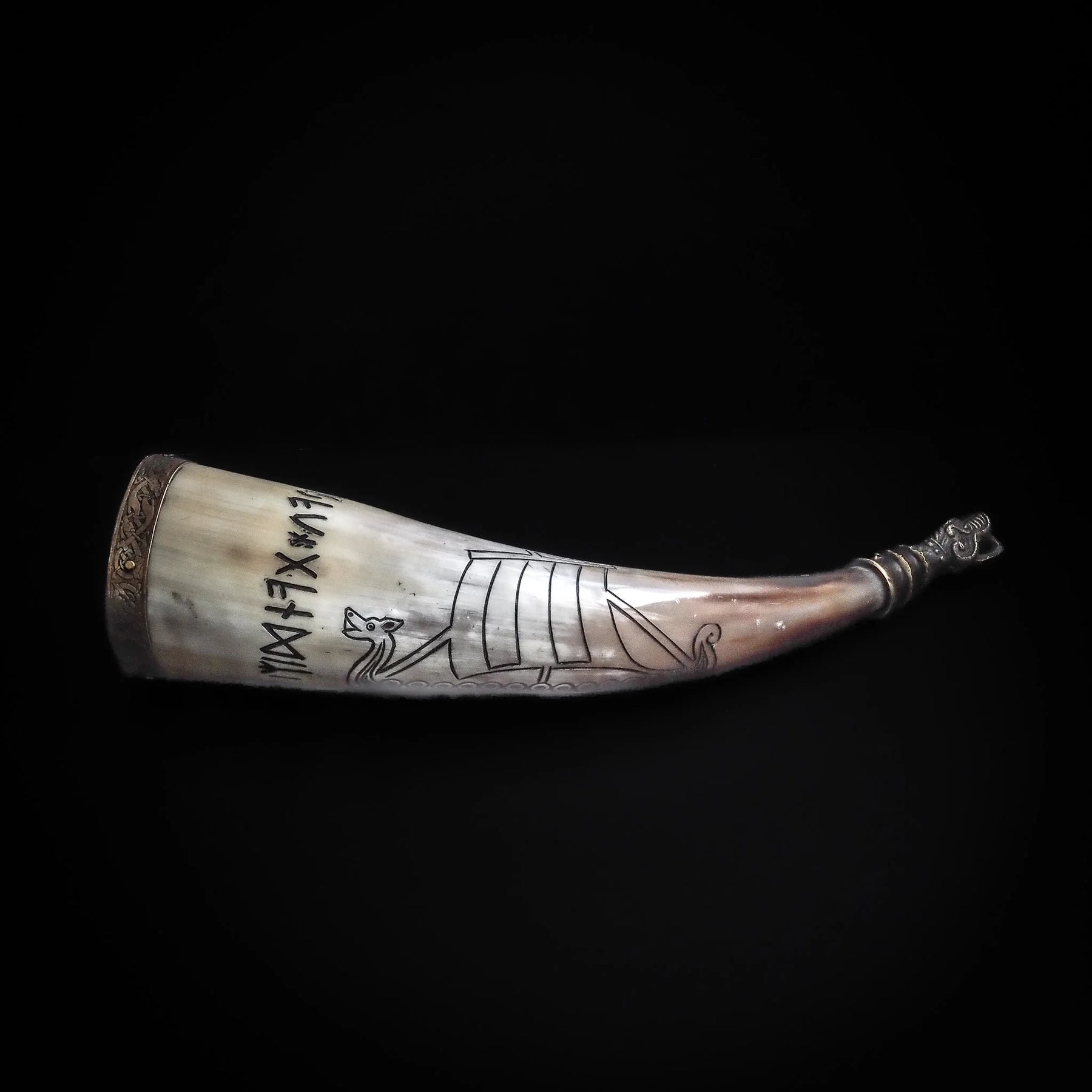 Dryckeshorn med organisk hornmaterial, graverad med korp och vikingaskepp. Mässingsbåge vid toppen med nordisk dekoration. Fotografi från sidan på svart bakgrund.