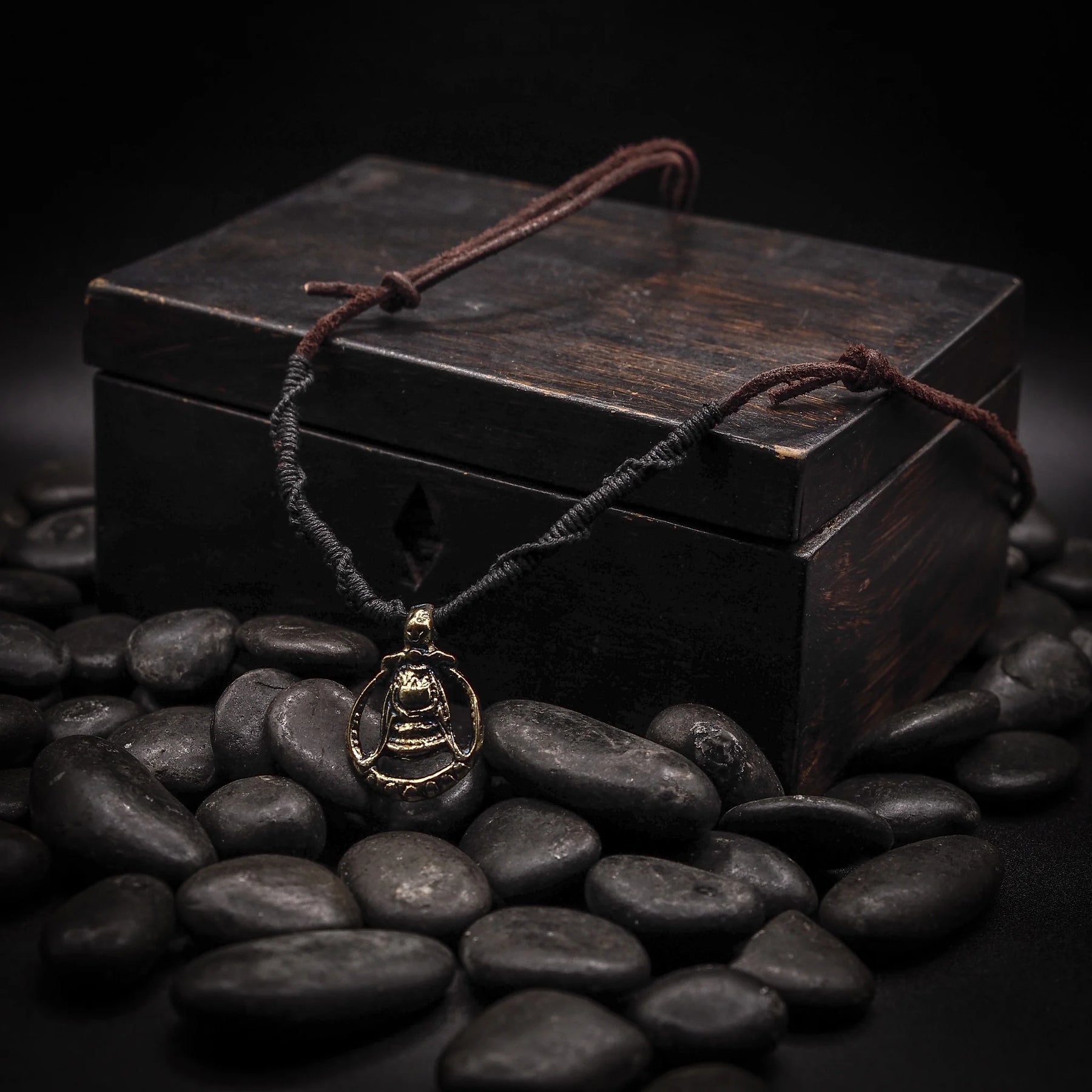 Ett silverhänge formadat som en Mammen-yxa med detaljerade mönster, hängande på läder- och naturfiber-snöre över en svart träkista omgiven av svarta stenar, fotograferat i studiomiljö.