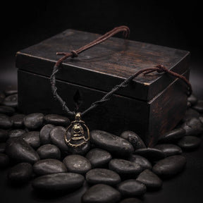 Ett silverhänge formadat som en Mammen-yxa med detaljerade mönster, hängande på läder- och naturfiber-snöre över en svart träkista omgiven av svarta stenar, fotograferat i studiomiljö.