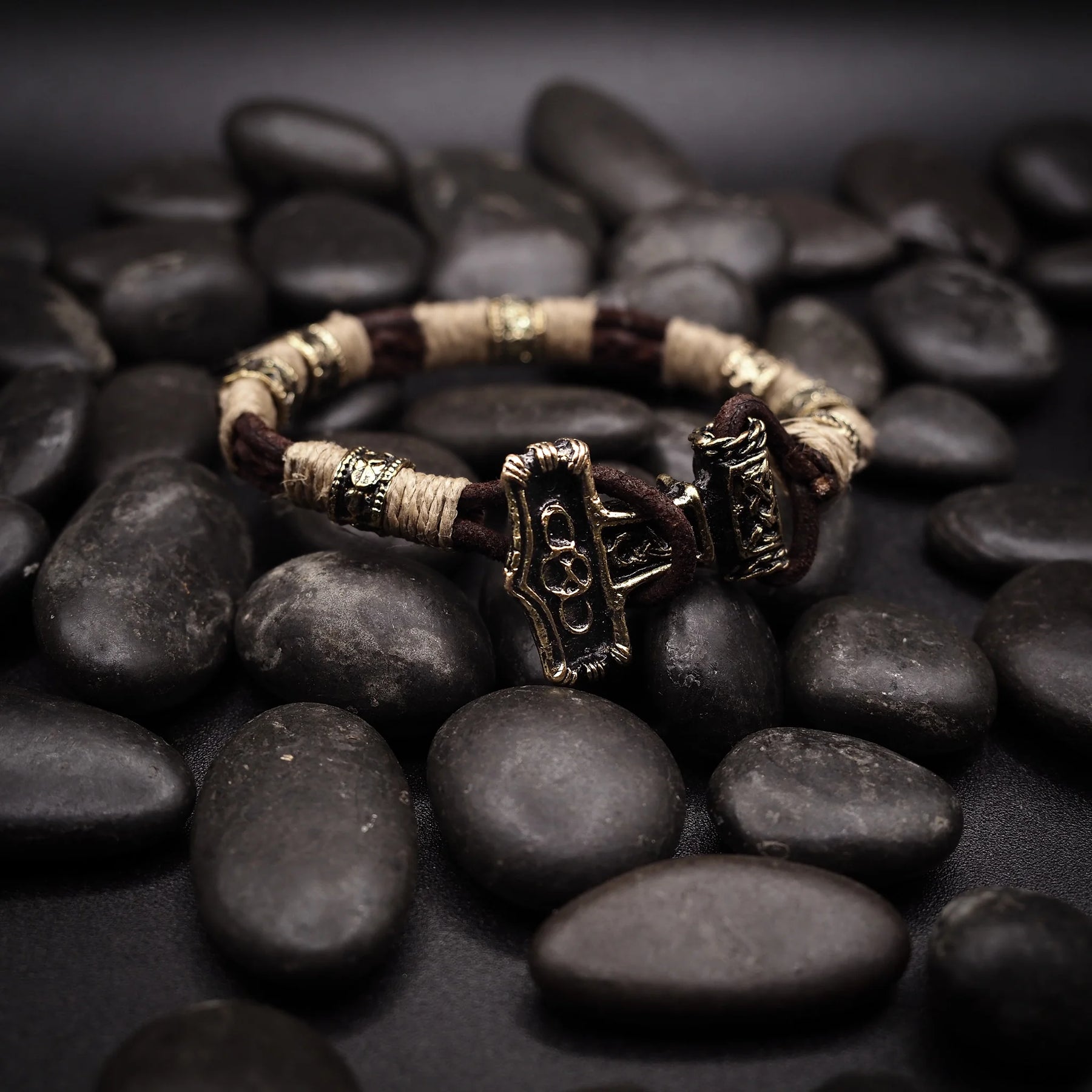 Vikingarmband på svart stenunderlag. Öppen armring med tre torshammare-anhängare graverade med vikingatida djurmotiv och geometriska mönster. Kräm-färgat rep och mässingsband skapar segmenterad design. Detaljerad mässingsarbete visar autentisk nordisk smyckestradition.
