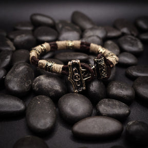Vikingarmband på svart stenunderlag. Öppen armring med tre torshammare-anhängare graverade med vikingatida djurmotiv och geometriska mönster. Kräm-färgat rep och mässingsband skapar segmenterad design. Detaljerad mässingsarbete visar autentisk nordisk smyckestradition.