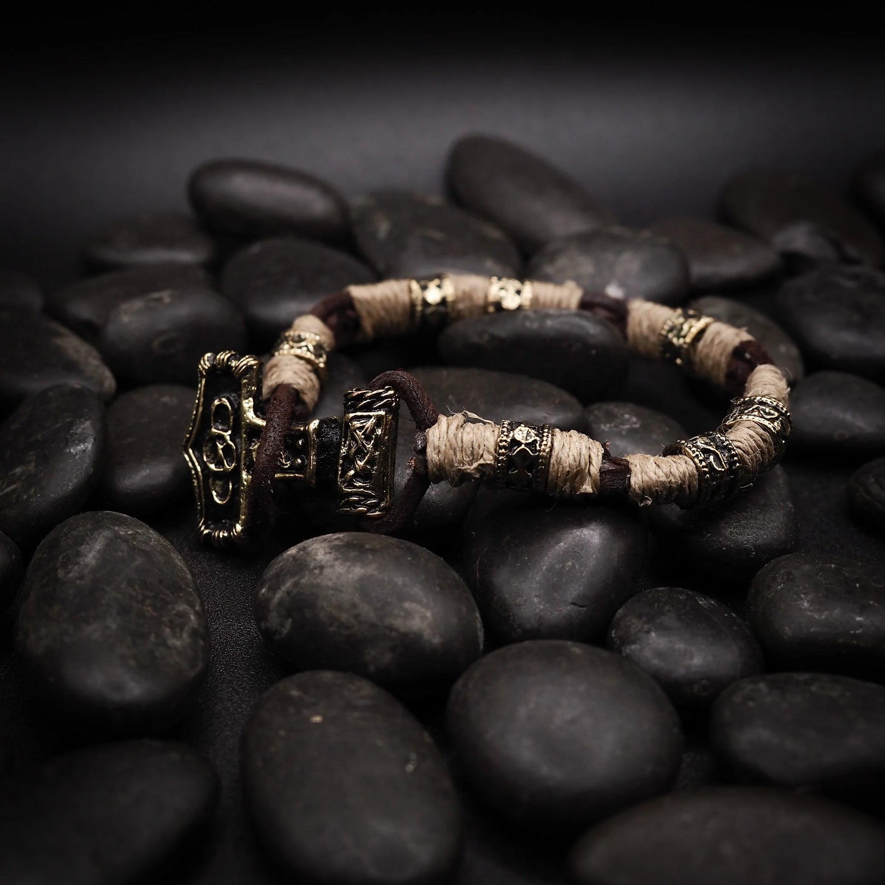 Vikingarmband på svarta stenar. Öppen manschettkonstruktion med två prydnadsändar i graverad mässing. Varje ände visar två Torshammare-anhängare med detaljerade vikingatida motiv inklusive djurhuvuden och geometriska mönster. Mittbandet är lindad med kräm-färgat rep och mässingsseparatorer.
