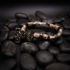 Vikingarmband på svarta stenar. Öppen manschettkonstruktion med två prydnadsändar i graverad mässing. Varje ände visar två Torshammare-anhängare med detaljerade vikingatida motiv inklusive djurhuvuden och geometriska mönster. Mittbandet är lindad med kräm-färgat rep och mässingsseparatorer.