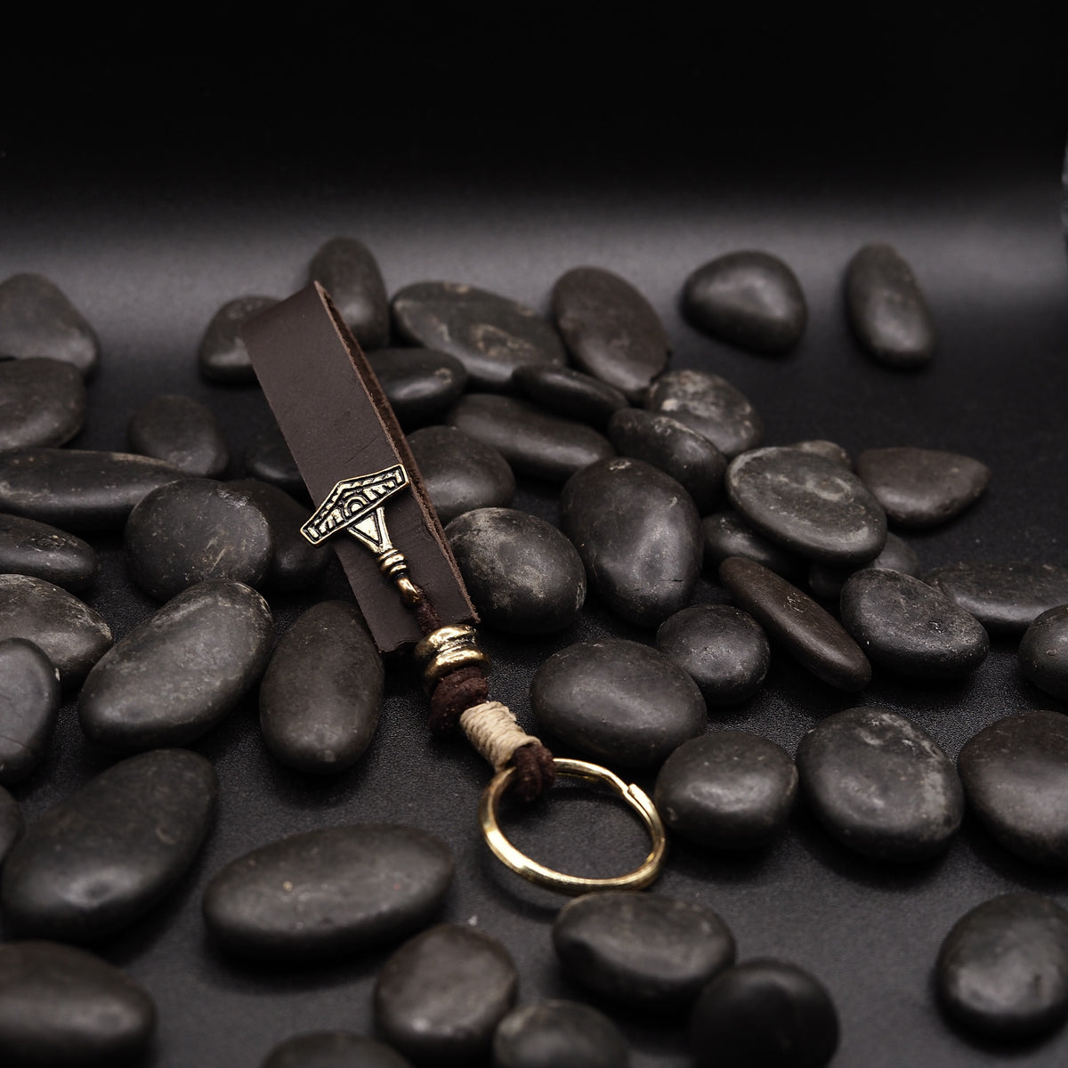 Nyckelring av lädre med liten Torshammare-amulett i brons, lagd på svarta stenar.