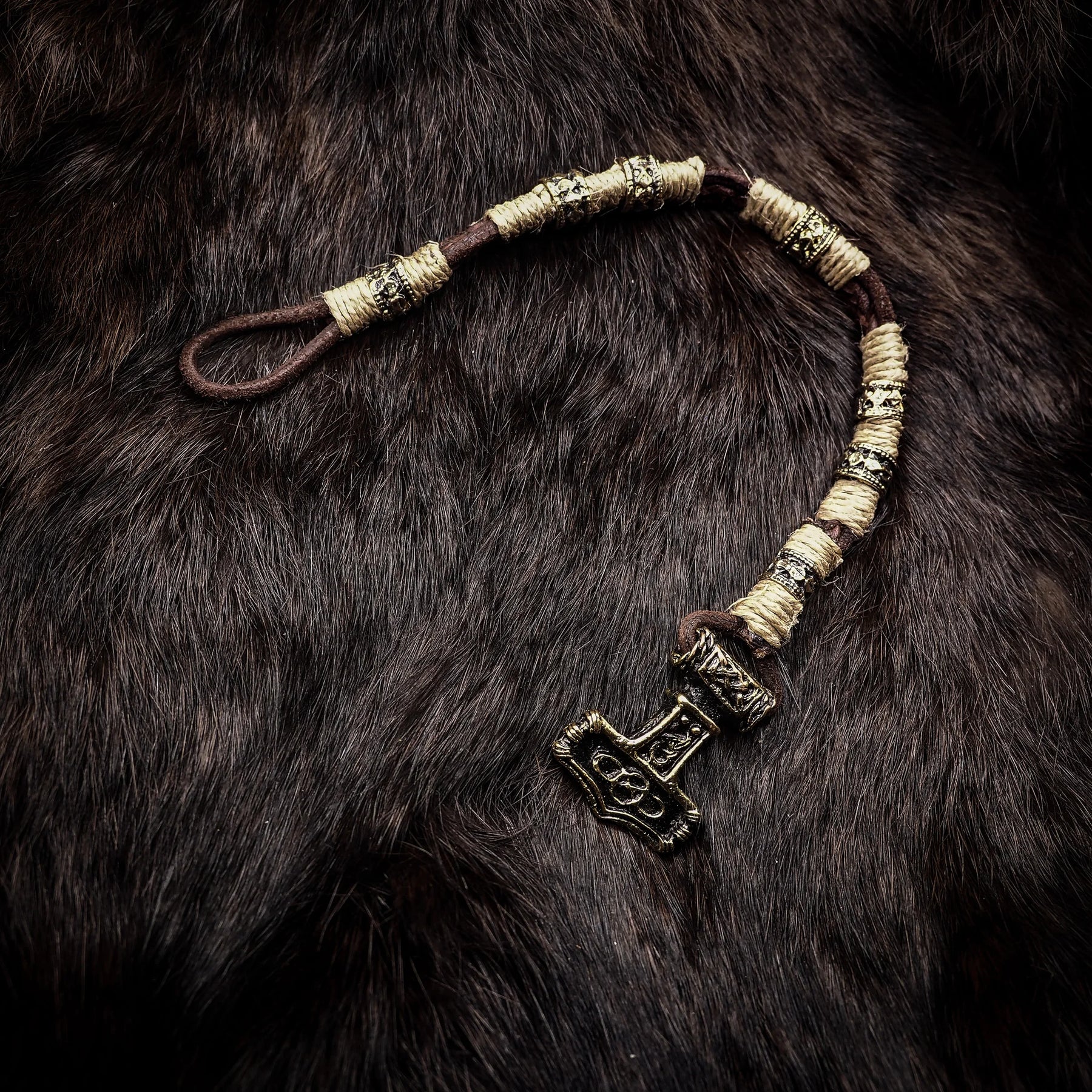Vikingarmband med torshammarfäste på mörkbrun päls. Armbandet har bredstaplade mässingssegment separerade av rottat rep i kräm och mörkbrunt. Torshammaranhängaren är ornamenterad med vikingatida geometriska mönster och fungerar som lås.