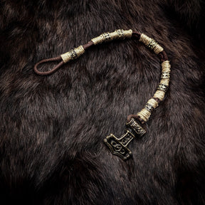 Vikingarmband med torshammarfäste på mörkbrun päls. Armbandet har bredstaplade mässingssegment separerade av rottat rep i kräm och mörkbrunt. Torshammaranhängaren är ornamenterad med vikingatida geometriska mönster och fungerar som lås.