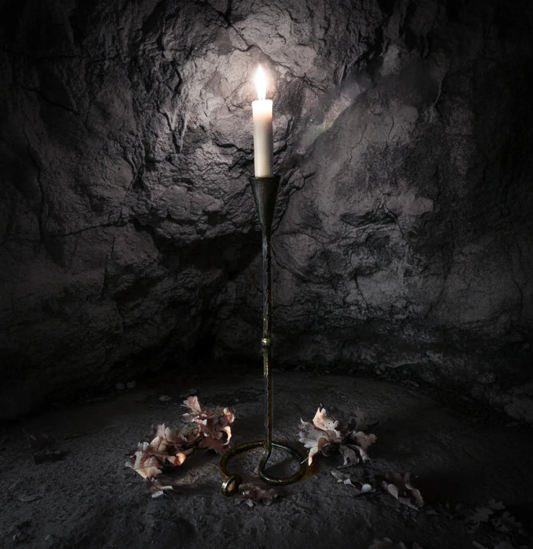 Lavatein ljusstake i hand-smiddat järn med tändstjärne som glödar inuti en mörk stenkavern. Dramatisk belysning framhäver den vikingska estetiken och det råa järnens finish.