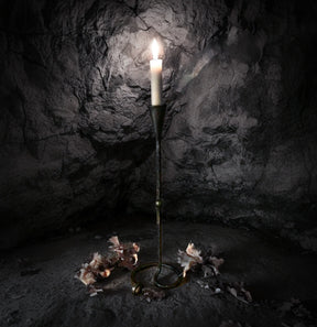 Lavatein ljusstake i hand-smiddat järn med tändstjärne som glödar inuti en mörk stenkavern. Dramatisk belysning framhäver den vikingska estetiken och det råa järnens finish.