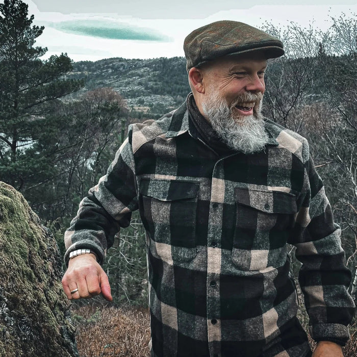 Skäggig man i rutig flanellskjorta och keps ler utomhus i skogsmiljö, livsstilsbild för Tor gåvoset