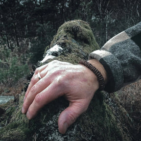 Brunt brokk-armband med väv- och flätmönster, skjutet på handled mot skogsbakgrund. Läderremmen är virad med metallknapp och loop-fäste.