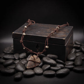 Pilspetshalsbandet liggande på mörkträ med svarta polerade stenar omkring. Varmt beige-gul stenspets lindad med mörkt snöre och med små benärlor längs två läderband. Studiofotografi med professionell belysning och skarp fokus på anhänget.