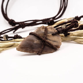 Pilspetshalsbandet i närbild på vit bakgrund. Beige-grå stenspets med naturliga kanter lindad i mörksnöre med läderband och benärlor. Vete och naturliga fibrer arrangerade konstnärligt omkring anhänget visar hantverk och neolitisk design.