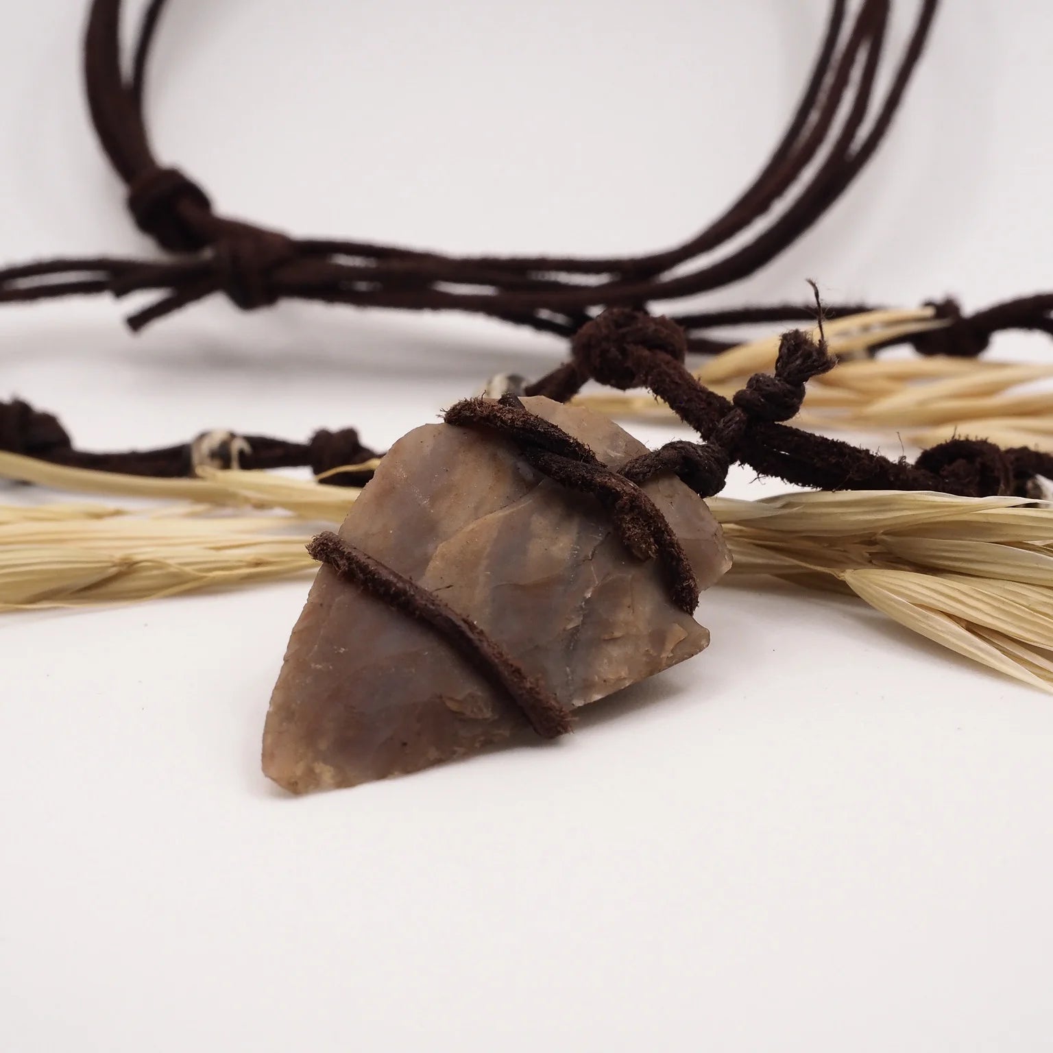 Pilspetshalsbandet på vit bakgrund presenterat minimalistiskt. Varm tan-beige stenspets med naturliga strieringar lindad i mörkbrunt snöre. Två läderband med benärlor för justerbar längd. Vete och naturligt material omkring visar hantverk och vikingaarv.