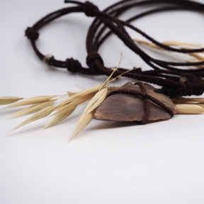 Närbild av pilspetsanhänget hållet i hand mot mörk bakgrund. Ljuscream-beige sten lindad med mörkbrunt snöre i geometriskt mönster. Stenens naturliga patina och neolitiska hantverk är tydligt synligt. Dramatisk kontrast framhäver detalj och bindningsteknik.