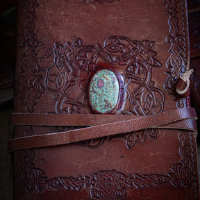 Handgjord läderbok Kristall i medium med rubin och fuxit-sten. Burgundibrunt präglat läder med vikingdekor, rödviolett sten med iriserande glans i metallfattning och bruna läderband.