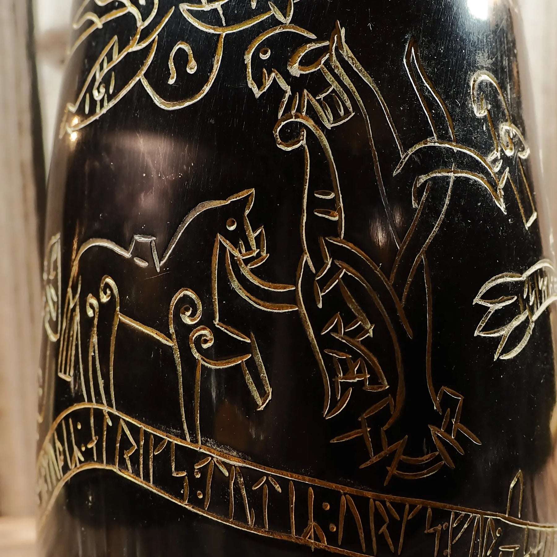 Närbild på graverad hornmugg med Sigurdsagan-detaljer. Fina snida av nordiska krigare, drakar och runor. Guldbetonande gravering på mörk horn visar högt hantverk.