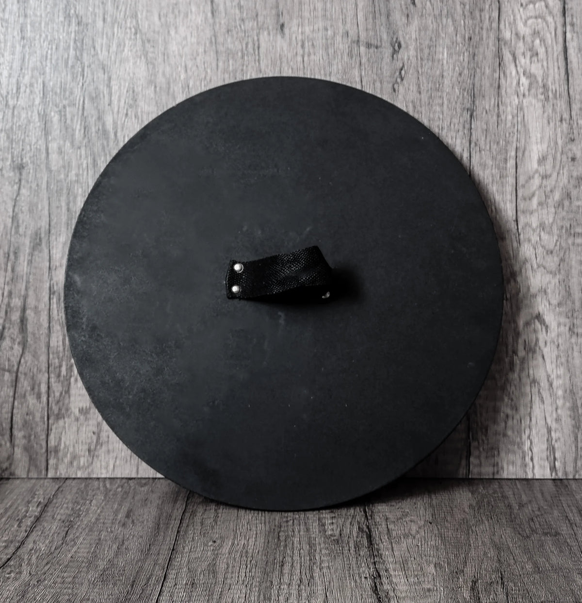Baksida av grön vikingsköld för barn. Svart MDF-yta med nylonhandtag i mitten. Skölden visar sitt tjocka rundade profil mot grå träbakgrund.