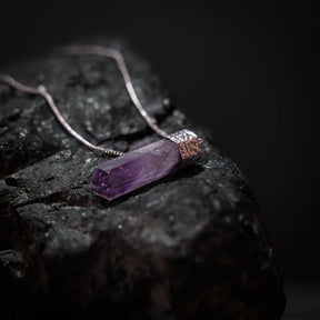 Lila ametistkristallhänge med silverfärgat mässingsfäste på kedja, placerat på mörkgrå sten