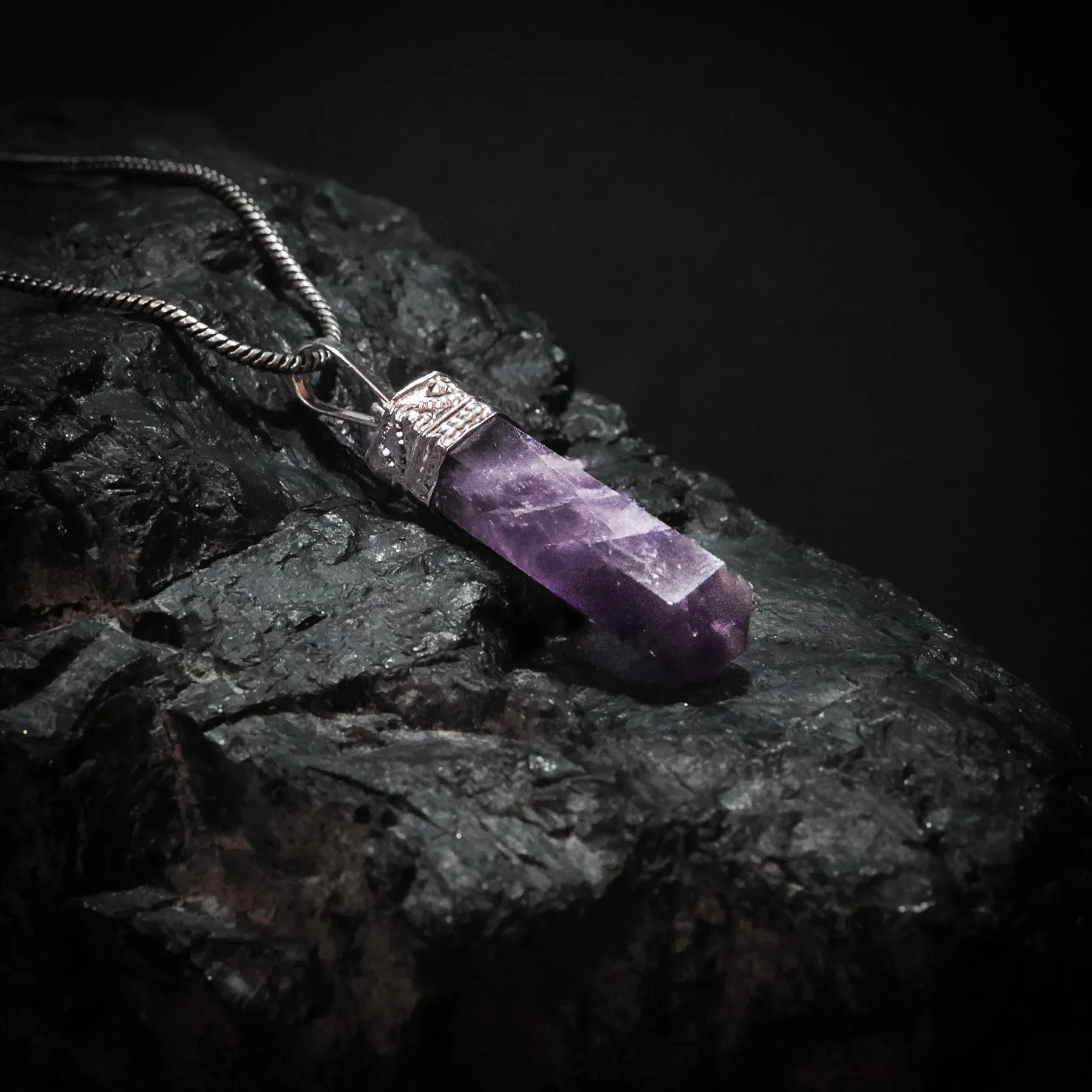 Ametisthänge i rå kristallform med silverfärgat mässingsfäste, upphängd på silvrig kedja mot mörk bakgrund