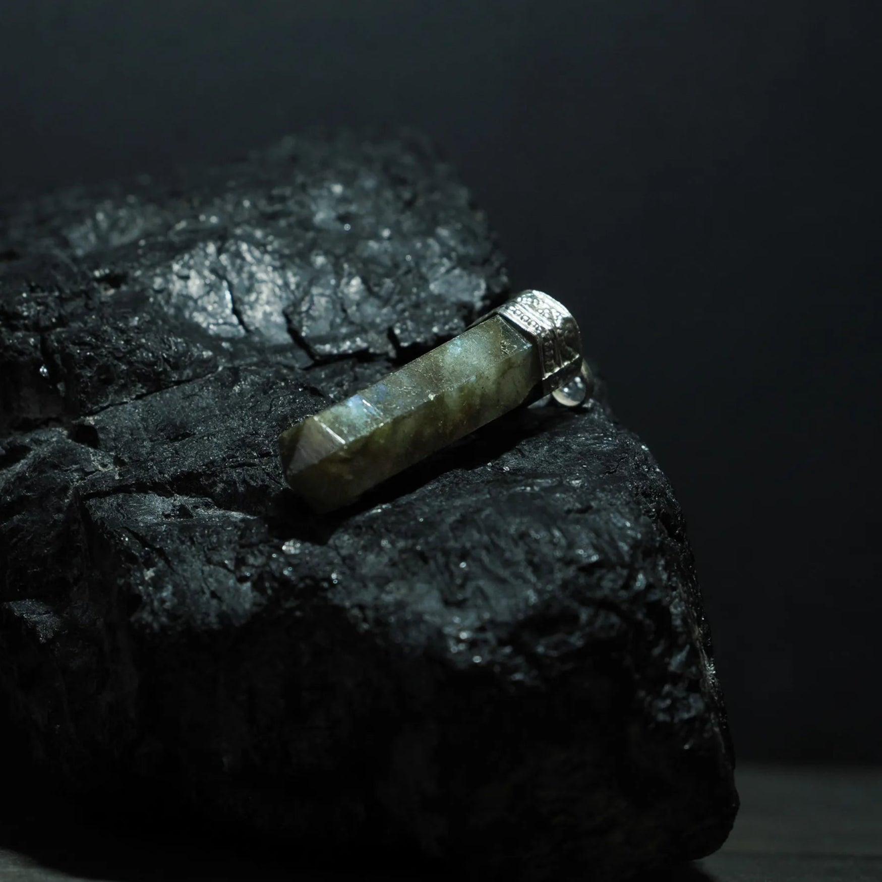 Labradorit-kristallhänge på mörkblå snöre, hängande mot mörk bakgrund. Stenens iriserade ljusblixtar dramatiskt belyst. Metallfattning med smyckesdetaljer. Lifestyle-produktfoto för halsbandspresentering.