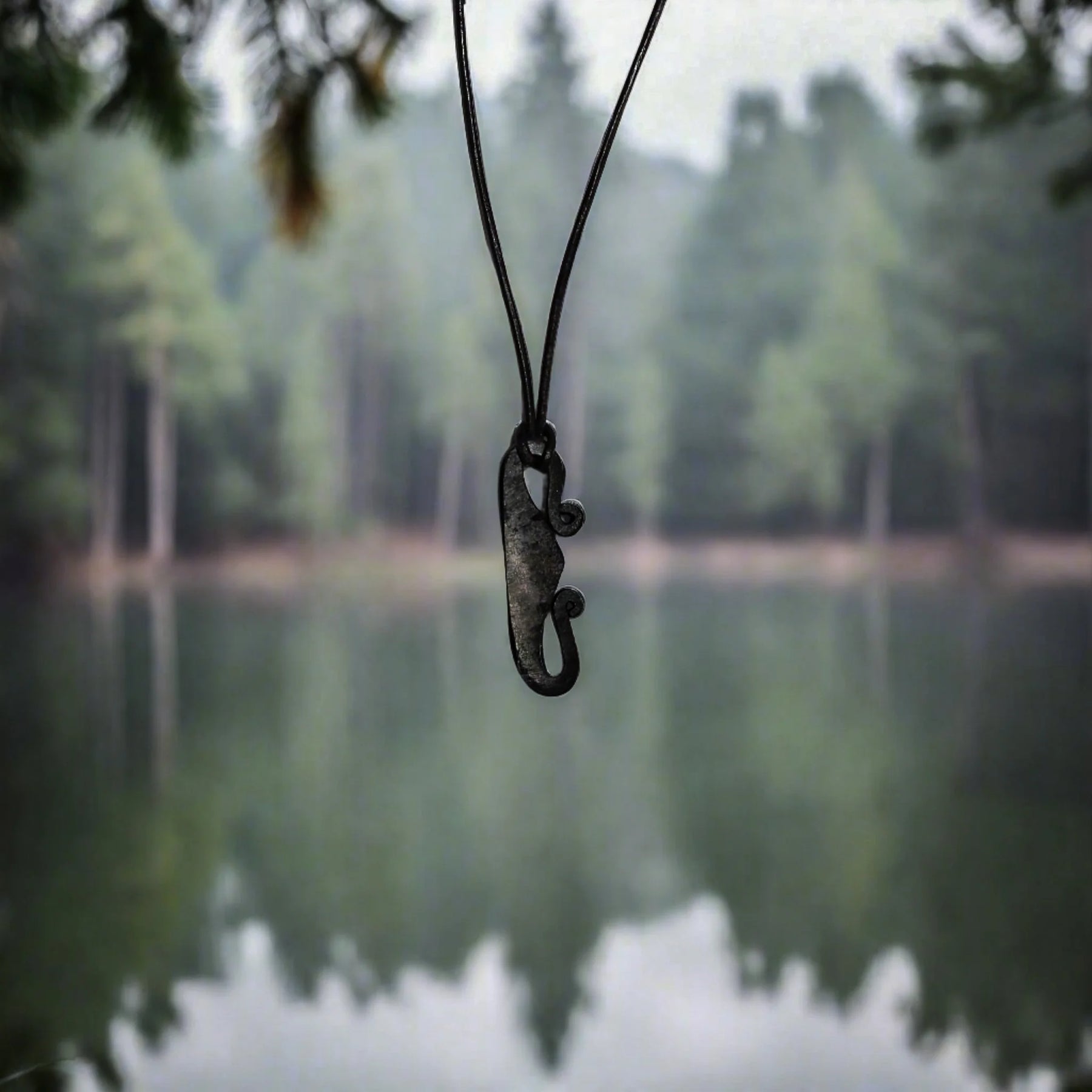Eldstålshalsband med järnpendant formad som Viking-verktyg, hängande på svarta snören mot en suddig skogssjöbakgrund