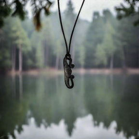 Eldstålshalsband med järnpendant formad som Viking-verktyg, hängande på svarta snören mot en suddig skogssjöbakgrund