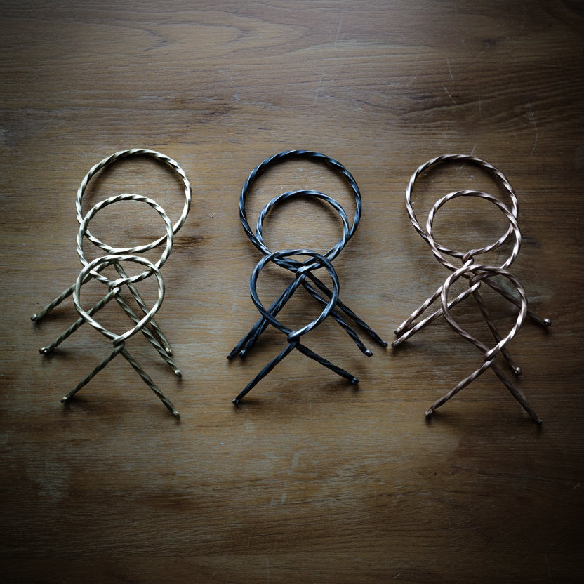 Tre hornställ i olika metallfinish (silver, svart, och koppar) arrangerade på rustik trävita bakgrund. Visar de vridna designen och metallets karakteristiska glans.